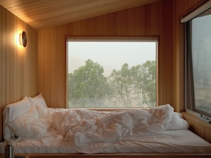 Een bed met witte lakens, een houten plafond, een raam met uitzicht op bomen en een wandlamp.