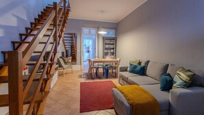 A modern living room with a wooden staircase, a grey sofa, a dining table with chairs, and a kitchen area in the background. - GuestReady - Casa Irmãs (Ponta Delgada)