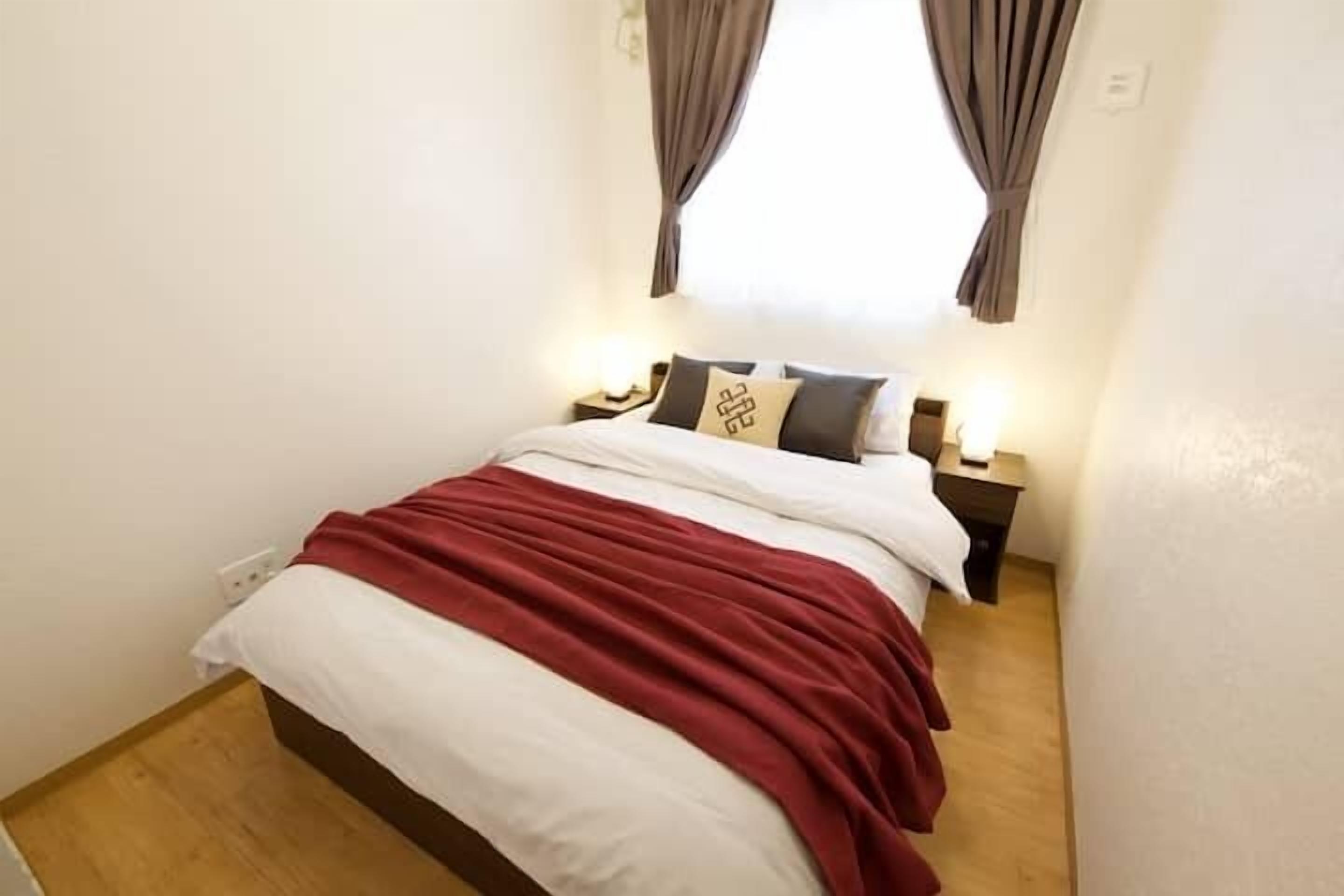 A bedroom with a bed, two bedside tables, a nightstand, a window with curtains, and a wall socket.