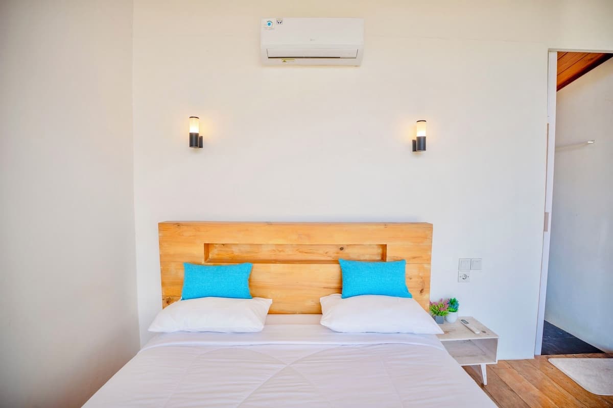 A bedroom with a wooden headboard, two white pillows, a small bedside table with a plant, and an air conditioning unit above the bed.