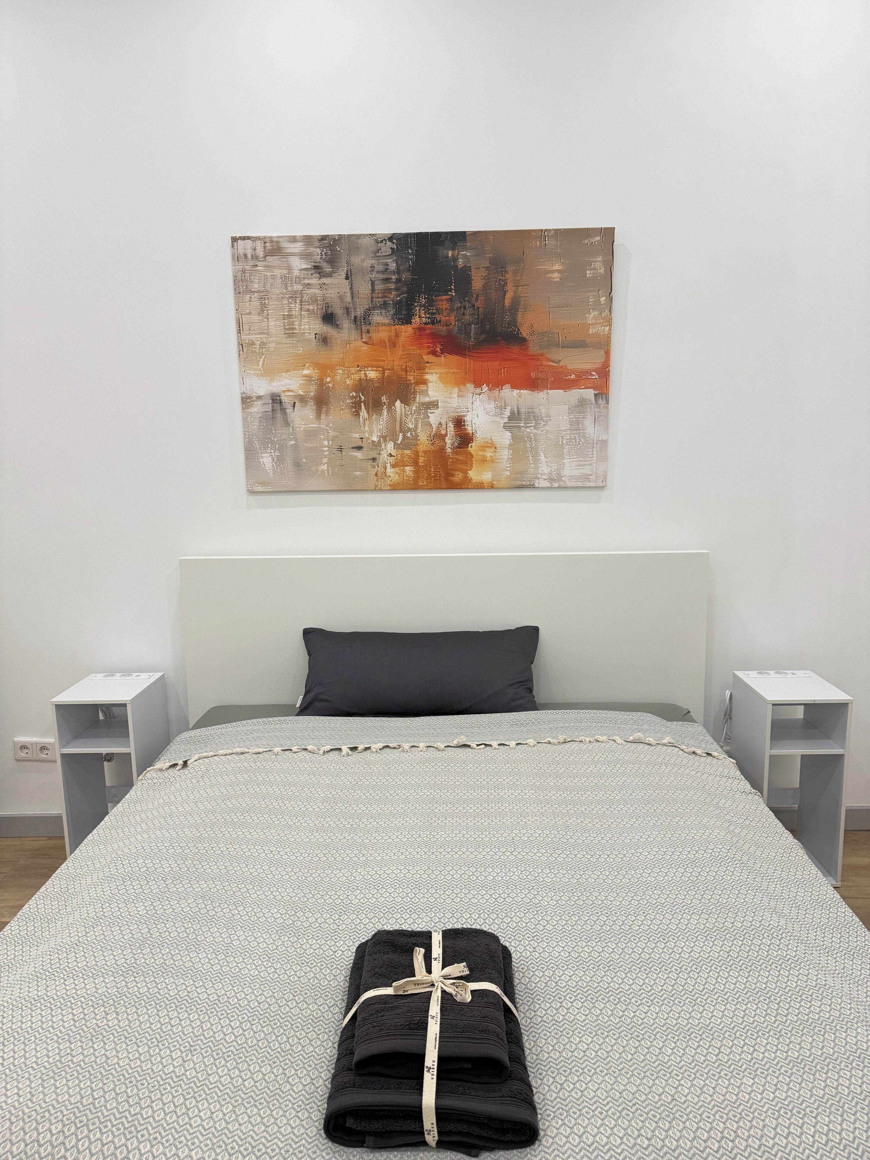 Ein ordentlich bezogenes Bett mit einer gemusterten Tagesdecke, einem dunklen Kopfkissen und einem gefalteten Handtuch mit Schleife.