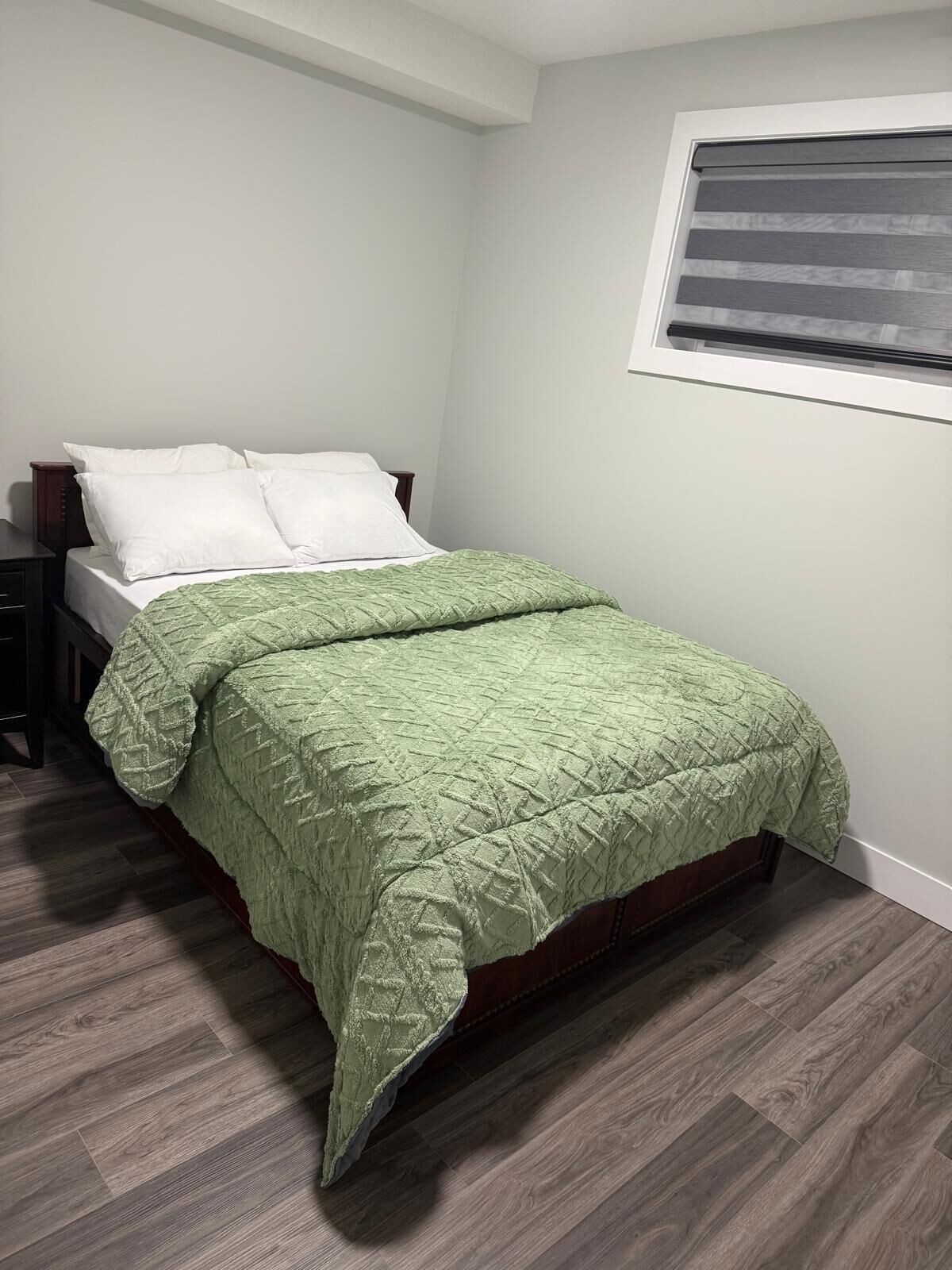 A bedroom with a bed, a nightstand, a window with blinds, and wooden flooring.