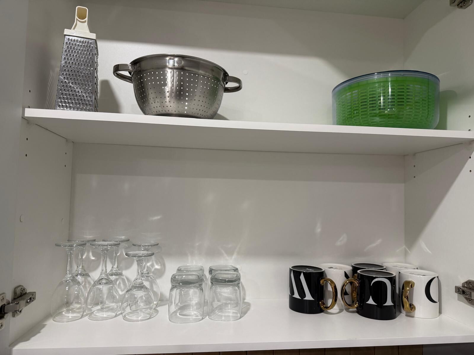 A kitchen shelf with a colander, a grater, and a green container. Below, there are wine glasses, jars, and mugs.