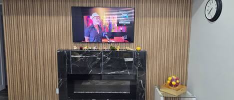 Una sala moderna con televisor de pantalla plana montado en una pared de madera, chimenea de mármol y una pequeña mesa con fruta.