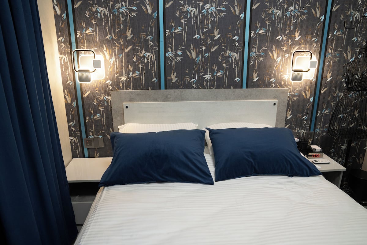 A hotel room with a bed, two bedside tables, a nightstand, and a wall with floral wallpaper.
