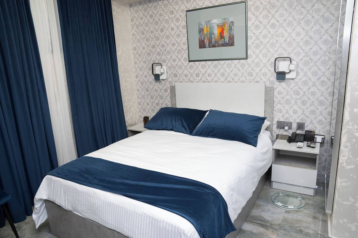 A neatly made bed with a white and blue bedspread, a white nightstand with a phone and a small table, and a wall-mounted light fixture.