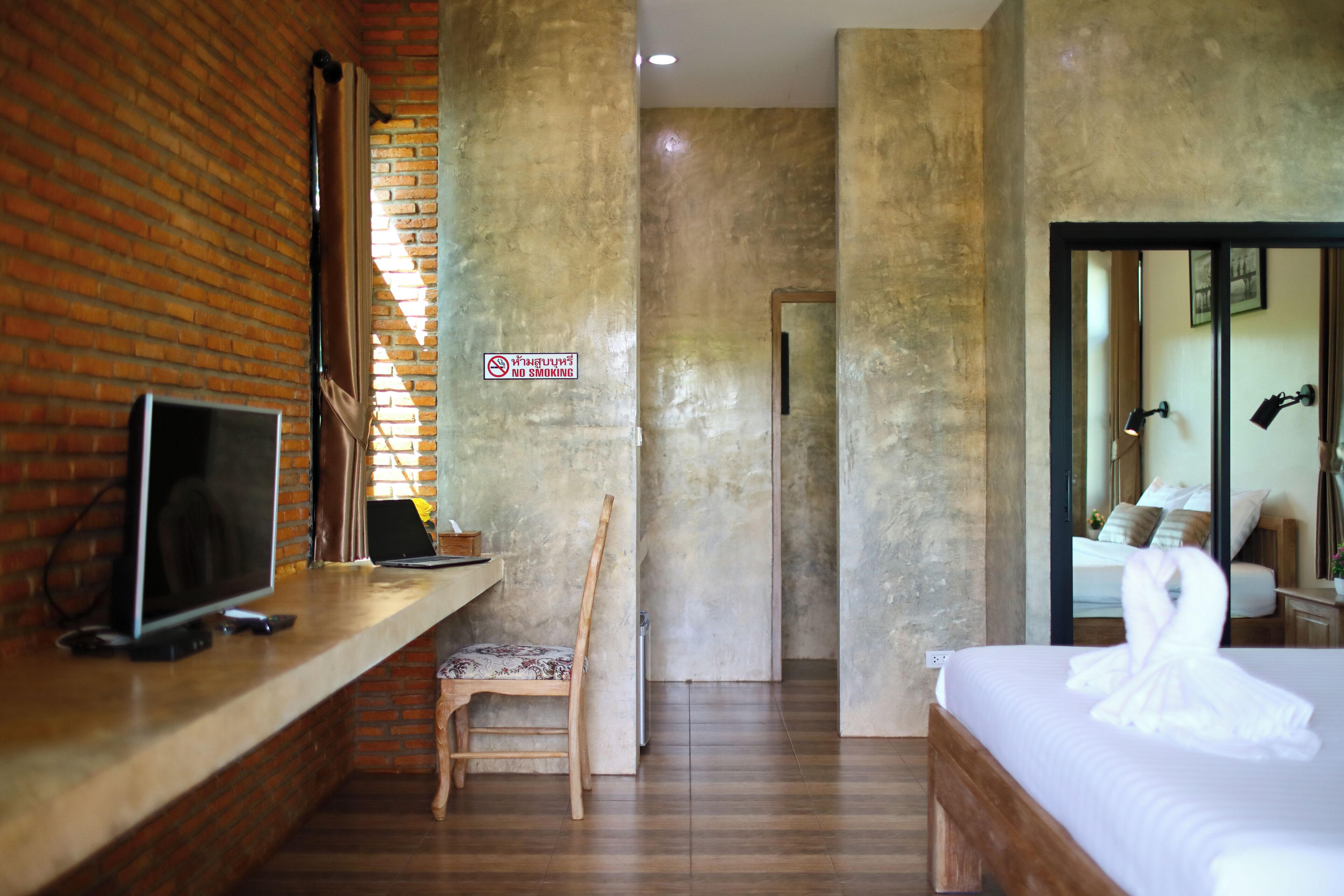 A hotel room with a brick wall, a television on a shelf, a wooden chair, a bed with a white towel, and a "No Smoking" sign.