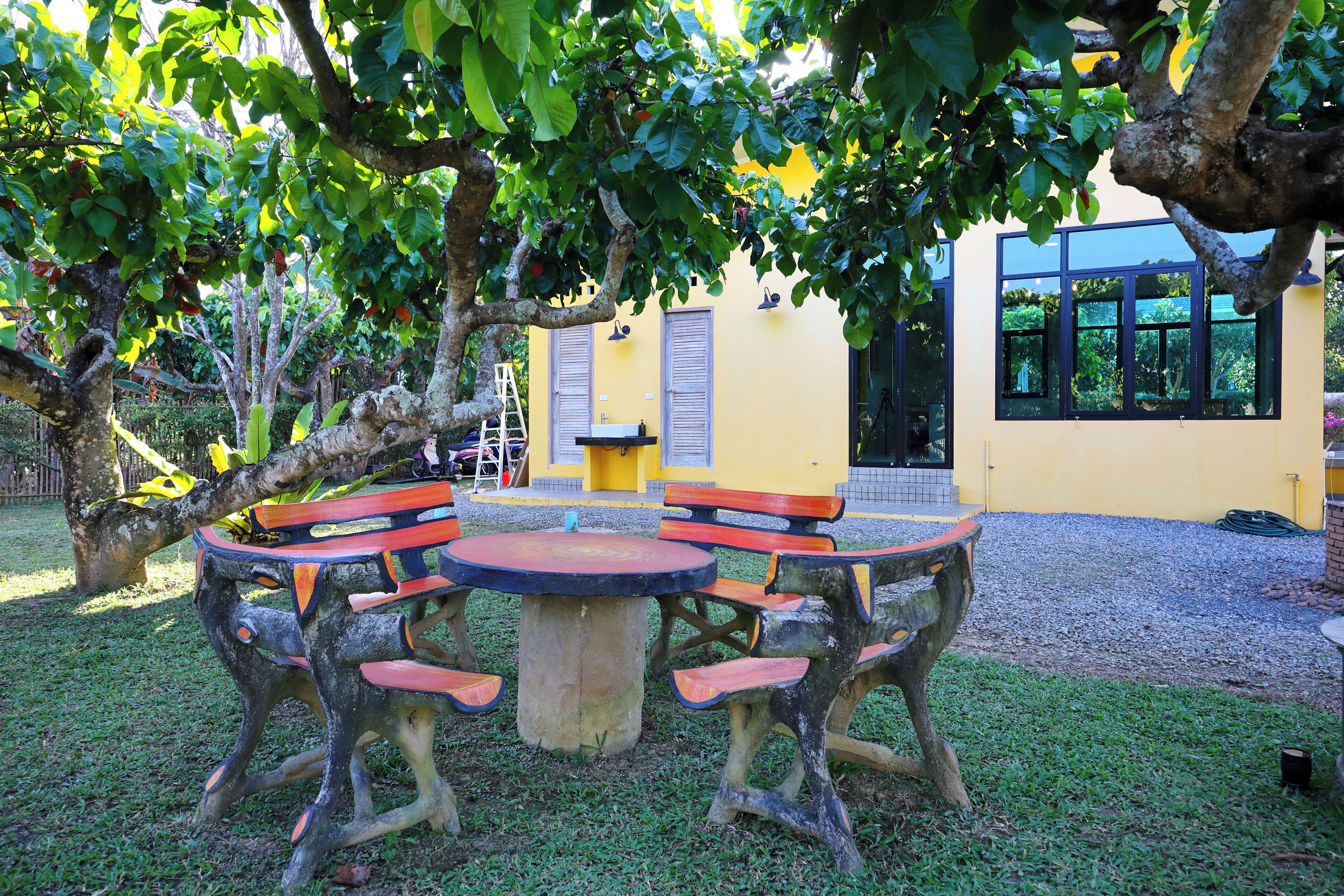 A round wooden table with benches, surrounded by trees, in a courtyard with a yellow building in the background.