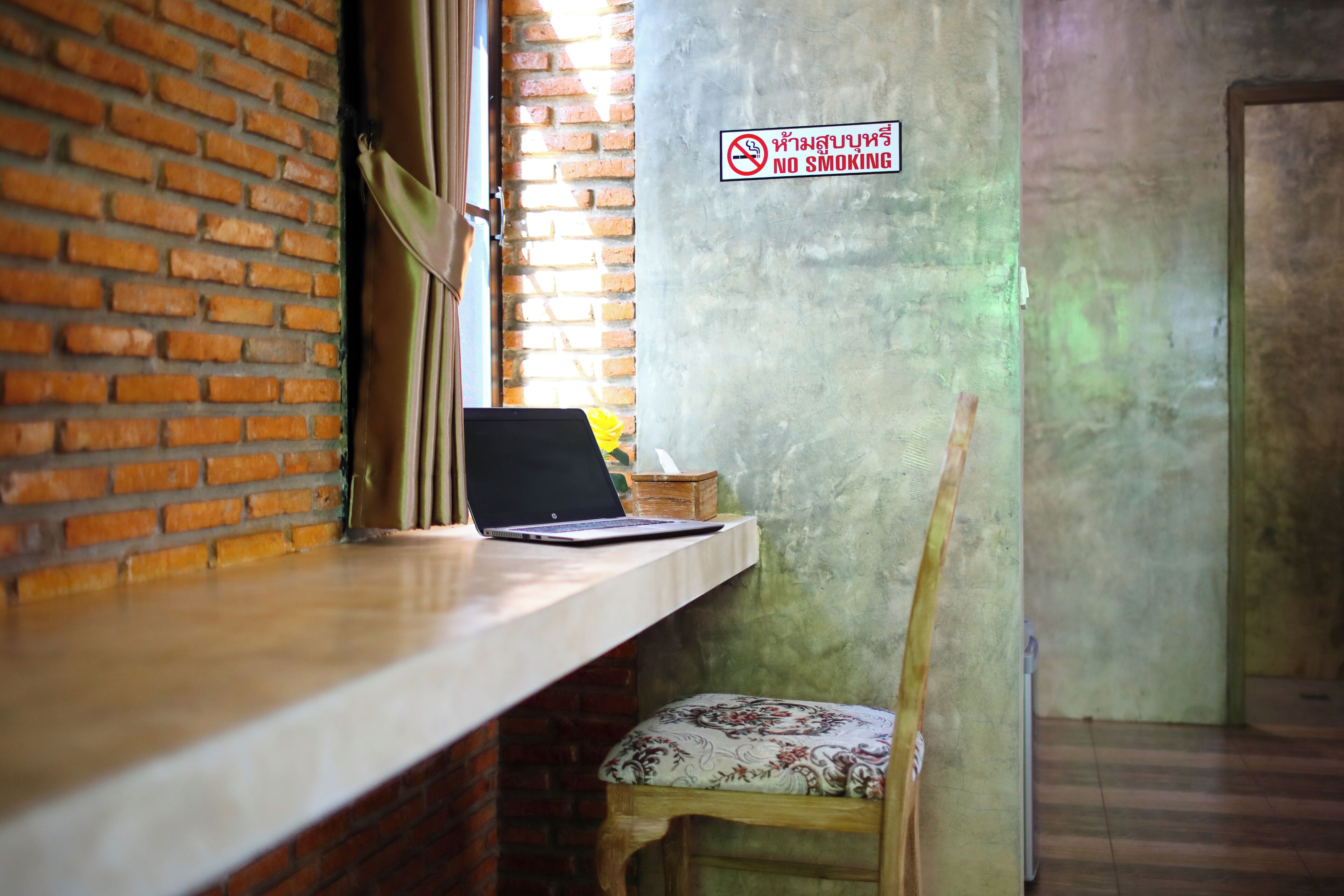 Una computadora portátil sobre un escritorio de madera con un pared de ladrillo y un cartel de 'No Smoking'.