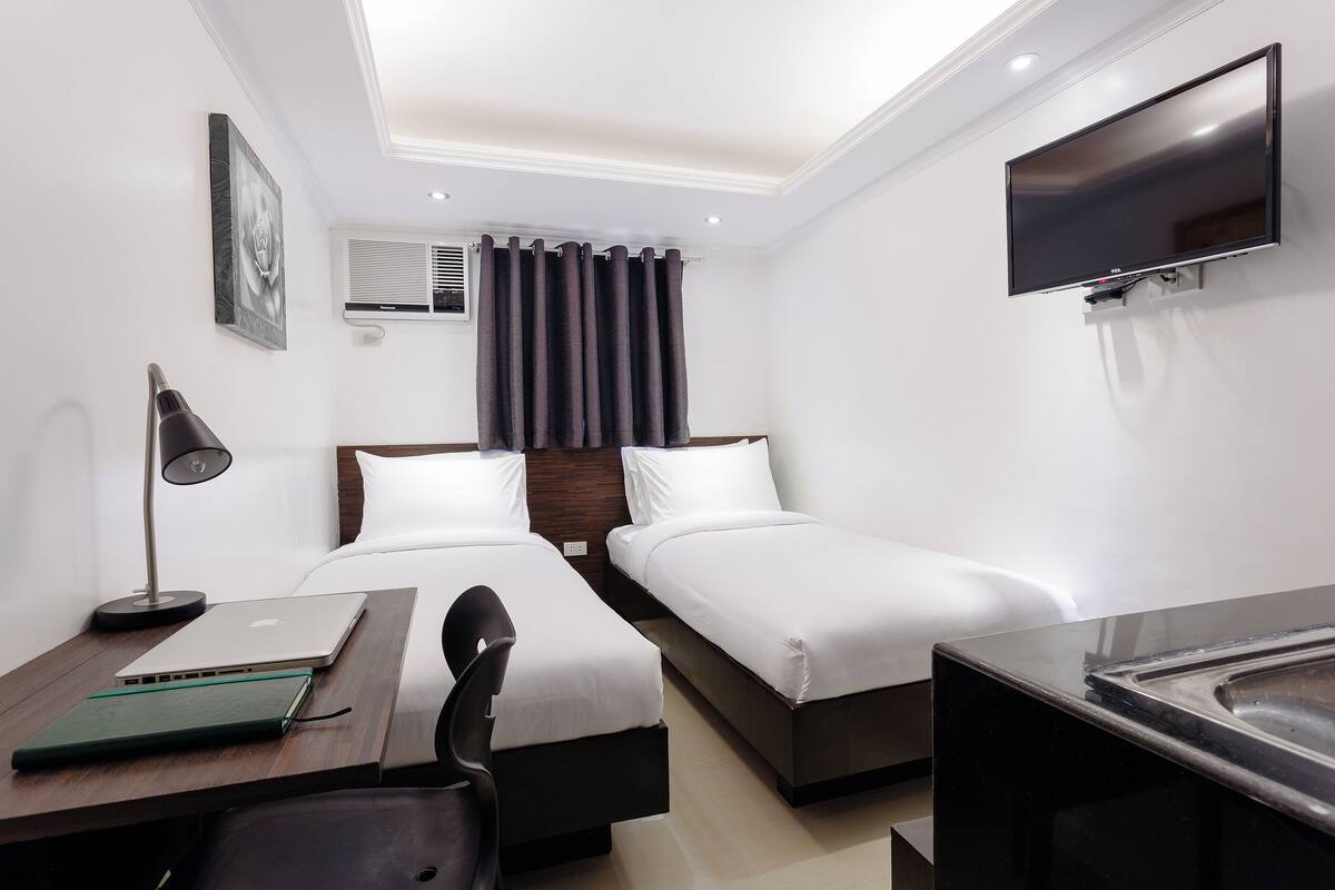 A hotel room with two single beds, a desk with a laptop, a chair, a TV mounted on the wall, and a window with curtains.