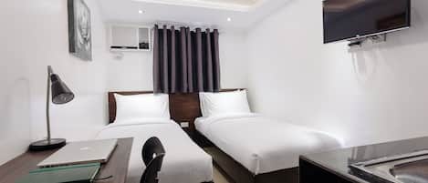 A hotel room with two single beds, a desk with a laptop, a chair, a TV mounted on the wall, and a window with curtains.