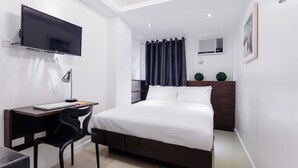 Een moderne slaapkamer met een bed, een bureau met een laptop, een aan de muur bevestigde televisie en een raam met gordijnen.