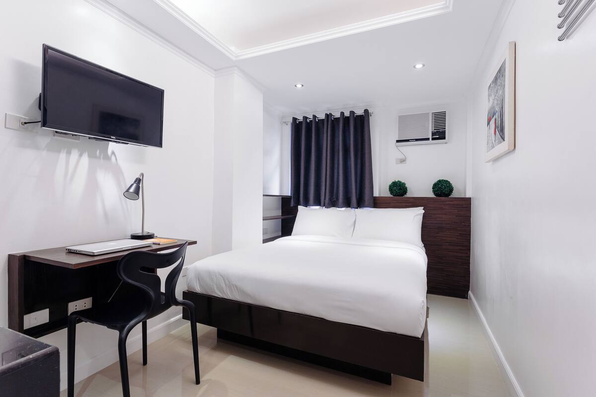 A modern bedroom with a bed, a desk with a laptop, a TV mounted on the wall, and a window with curtains.
