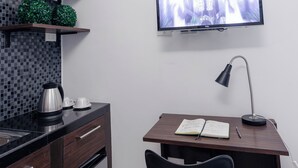 Een moderne keuken met een ingebouwde magnetron, waterkoker en koffieapparaat op het aanrecht. Er staat een houten bureau met een stoel en een televisie hangt aan de muur.