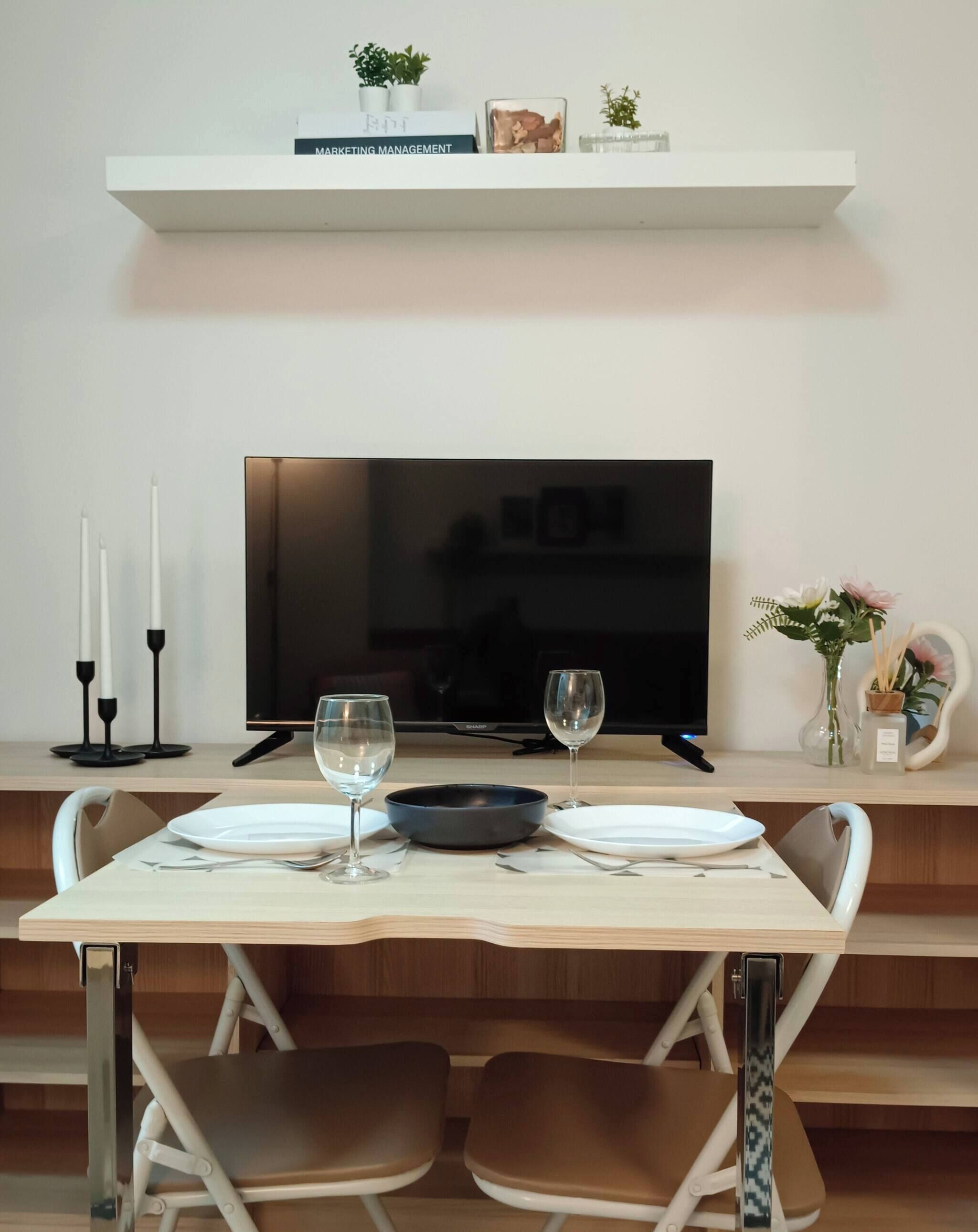 Un comedor moderno con una televisión de pantalla plana, una mesa blanca con dos sillas y un estante con un libro y objetos decorativos.