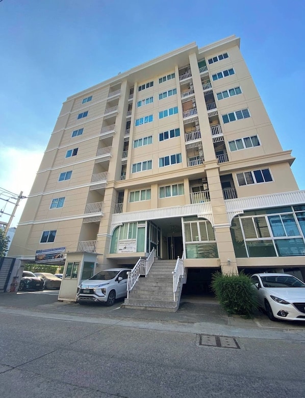 Un edificio de varios pisos con una entrada central, una escalera que conduce a la entrada y un estacionamiento con automóviles.