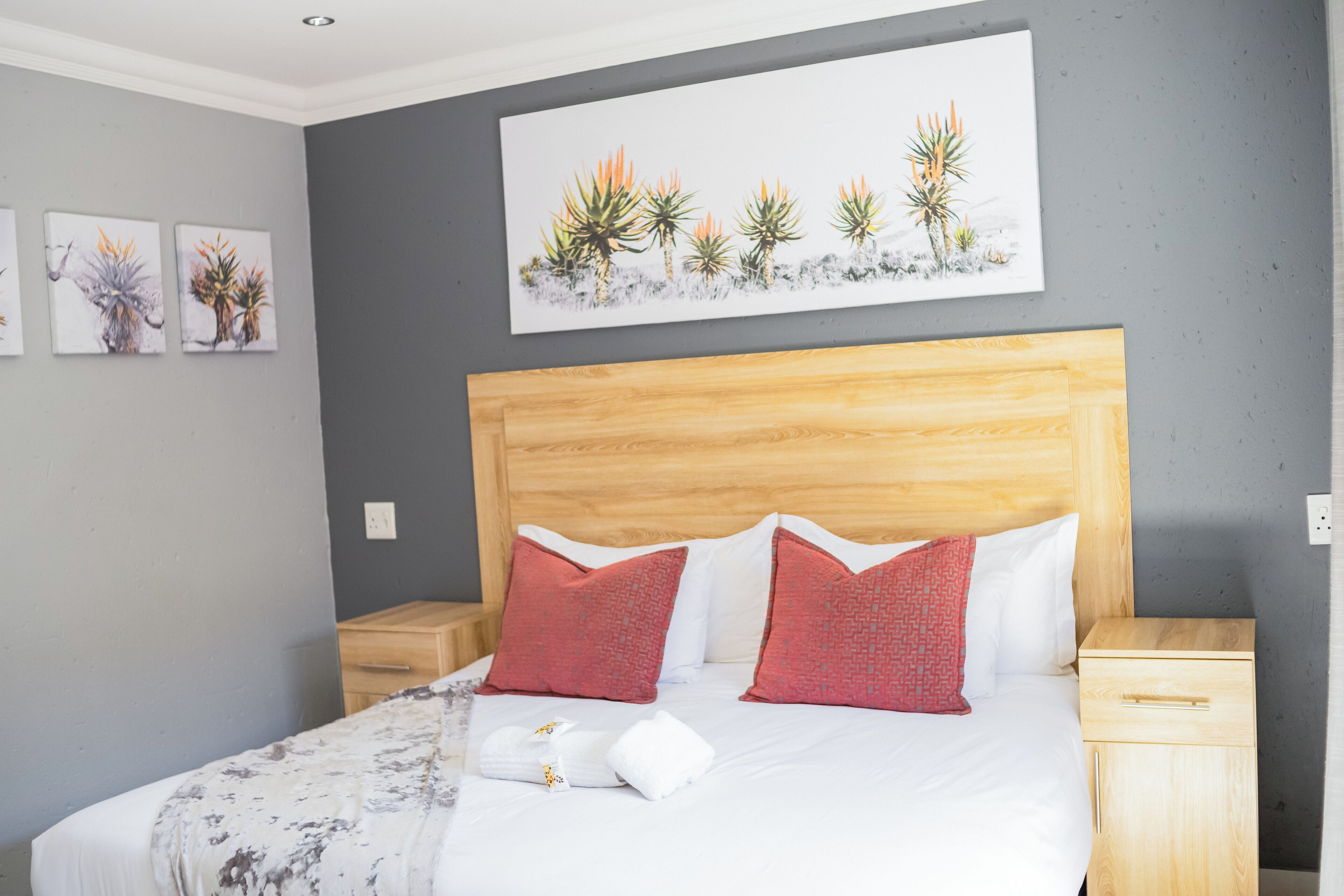 A neatly made bed with a wooden headboard, two red pillows, and a white blanket. There are two wooden nightstands with drawers, and a wall-mounted artwork of desert plants above the bed.