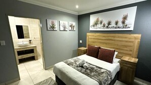 Un dormitorio con cama, mesitas de noche, cabecera de madera, baño con lavamanos y espejo, y obras de arte en la pared.
