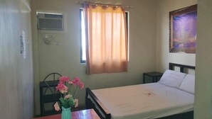 A small, single-bed room with a wooden headboard, a bedside table, a small table with a vase of flowers, a window with curtains, and a wall-mounted air conditioning unit. - WJV INN CASUNTINGAN (Mandaue)