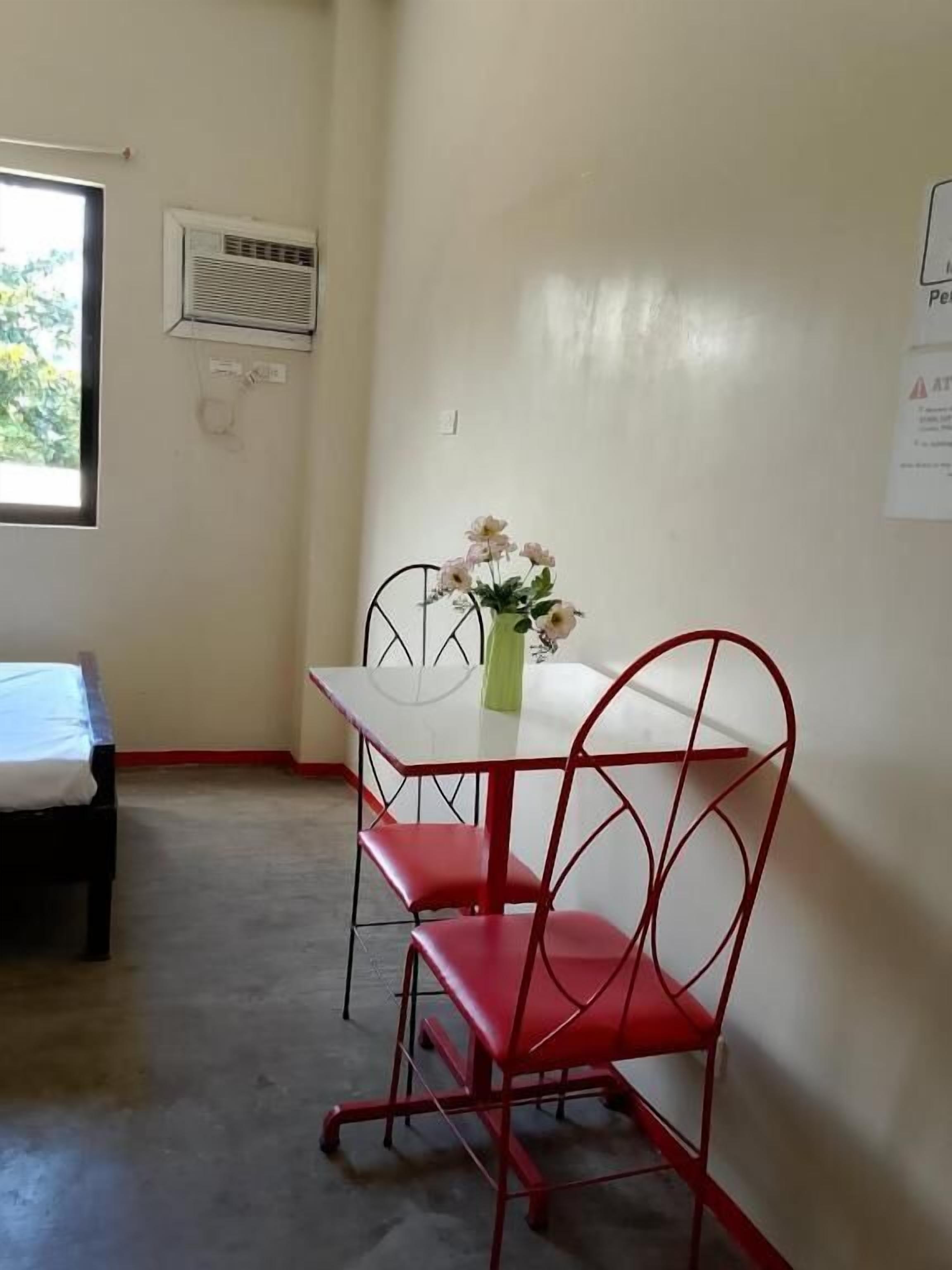 A small room with a white table and two red chairs, a window with blinds, and a wall-mounted air conditioning unit.