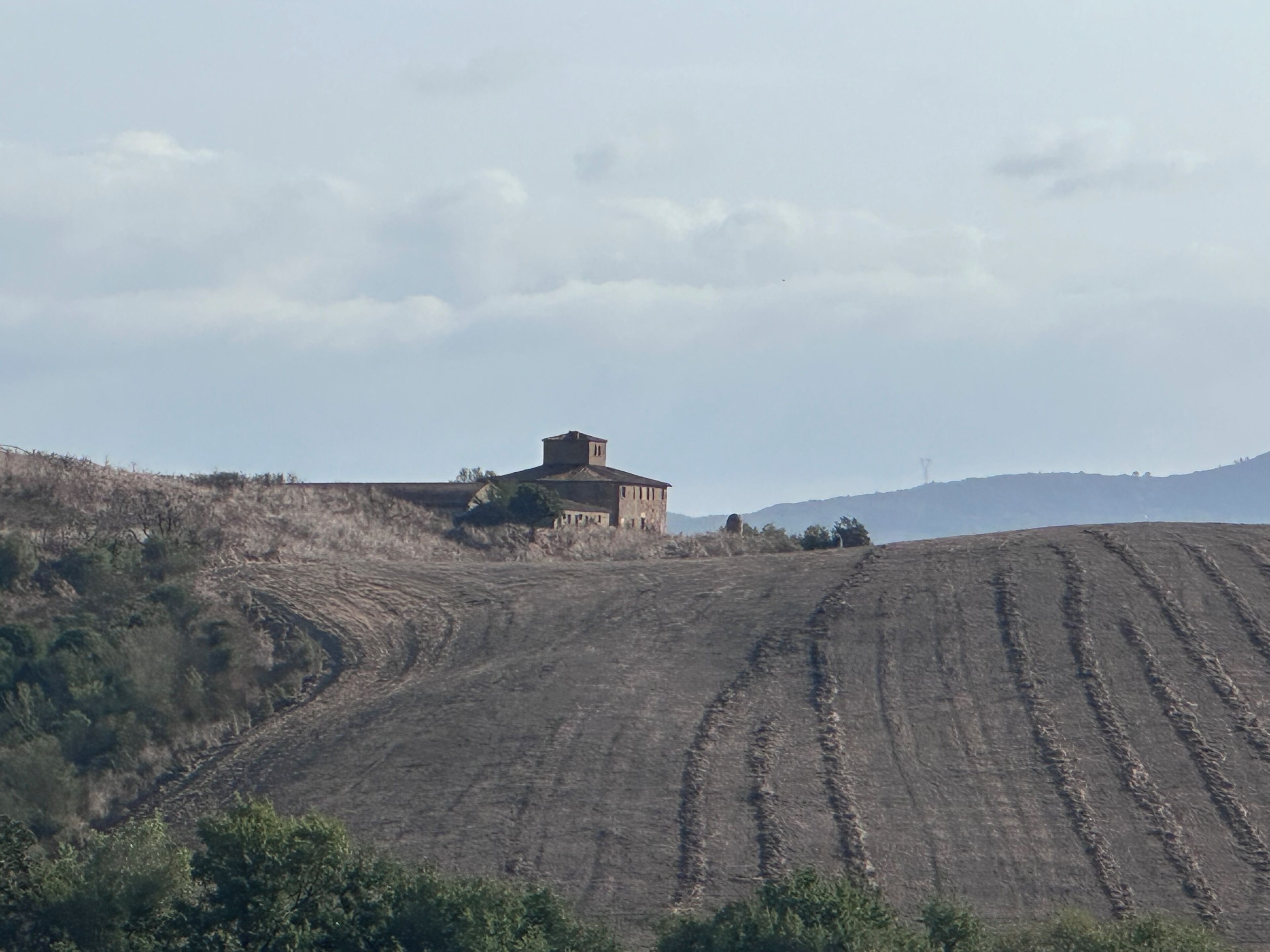 A rural landscape with a single building on a hill, surrounded by undulating terrain and sparse vegetation.
