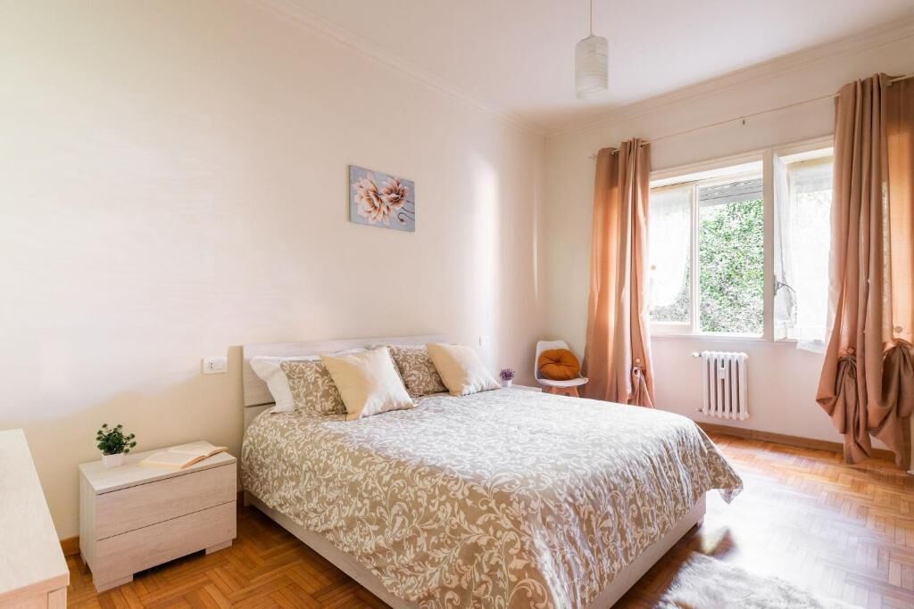 A bedroom with a large bed, a bedside table, a small chair, a window with curtains, and a radiator.