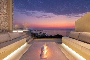 A modern outdoor seating area with a fire feature, overlooking a cityscape at sunset. - Monte Smith Villa Aether (C6F3+X4J Ialisos, Greece)