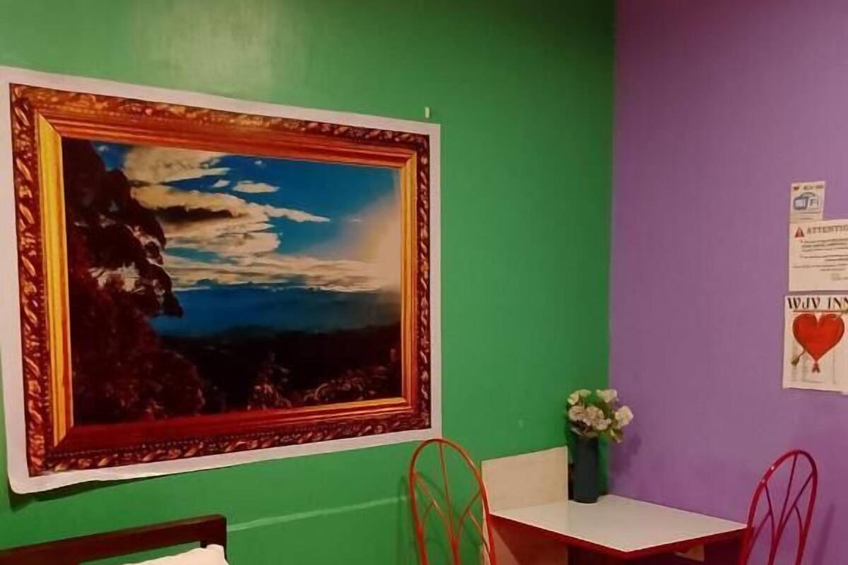 Una habitación con una cama, un escritorio y dos sillas rojas, un cuadro enmarcado y un jarrón con flores.