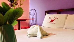 A neatly made bed with a pillow labeled "WJV- INN" and a heart symbol, a metal chair, and a potted plant with pink flowers. - WJV INN CANDUMAN (Mandaue)