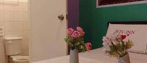 A hotel room with a bed, two vases with flowers, a painting on the wall, and a sign with the hotel name.