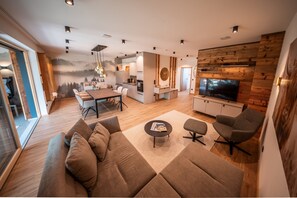 A modern living room with a large sofa, a coffee table, and a TV. There is a dining area with a table and chairs. The room has wooden flooring and a wooden accent wall.