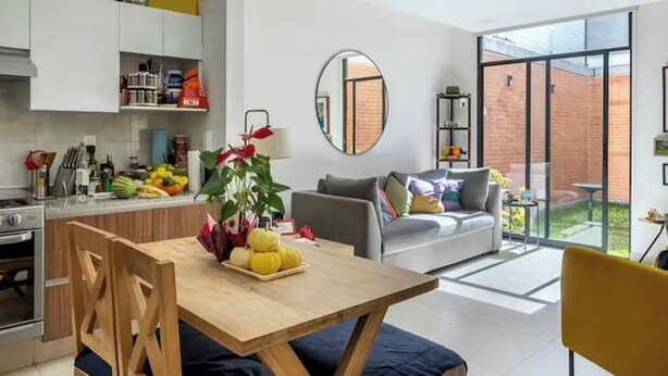 A modern kitchen with white cabinets, a dining table with a plant centerpiece, and a grey sofa with a view to a brick exterior. - Acogedor Departamento a 7 minutos de Coyoacán + Bonita azotea. (Ciudad de México)