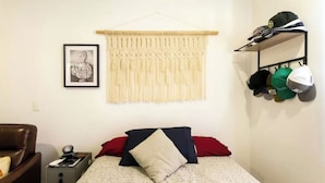 A bedroom with a bed, a framed picture, a macramé wall hanging, and a shelf with hats. - Acogedor Departamento a 7 minutos de Coyoacán + Bonita azotea. (Ciudad de México)