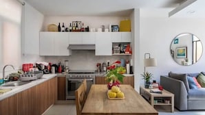 A modern kitchen with white cabinets, a wooden dining table with a fruit centerpiece, and a grey sofa in the living area. - Acogedor Departamento a 7 minutos de Coyoacán + Bonita azotea. (Ciudad de México)