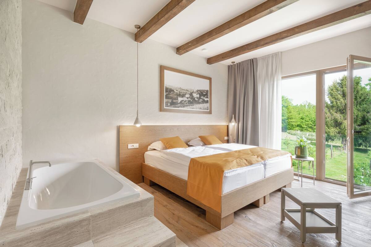 A modern bedroom with a large bathtub, a wooden bed, and a view of a garden through a sliding glass door.