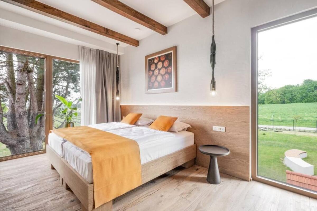 A bedroom with a large bed, a small side table, a hanging light fixture, and a view of a green landscape through a sliding glass door.