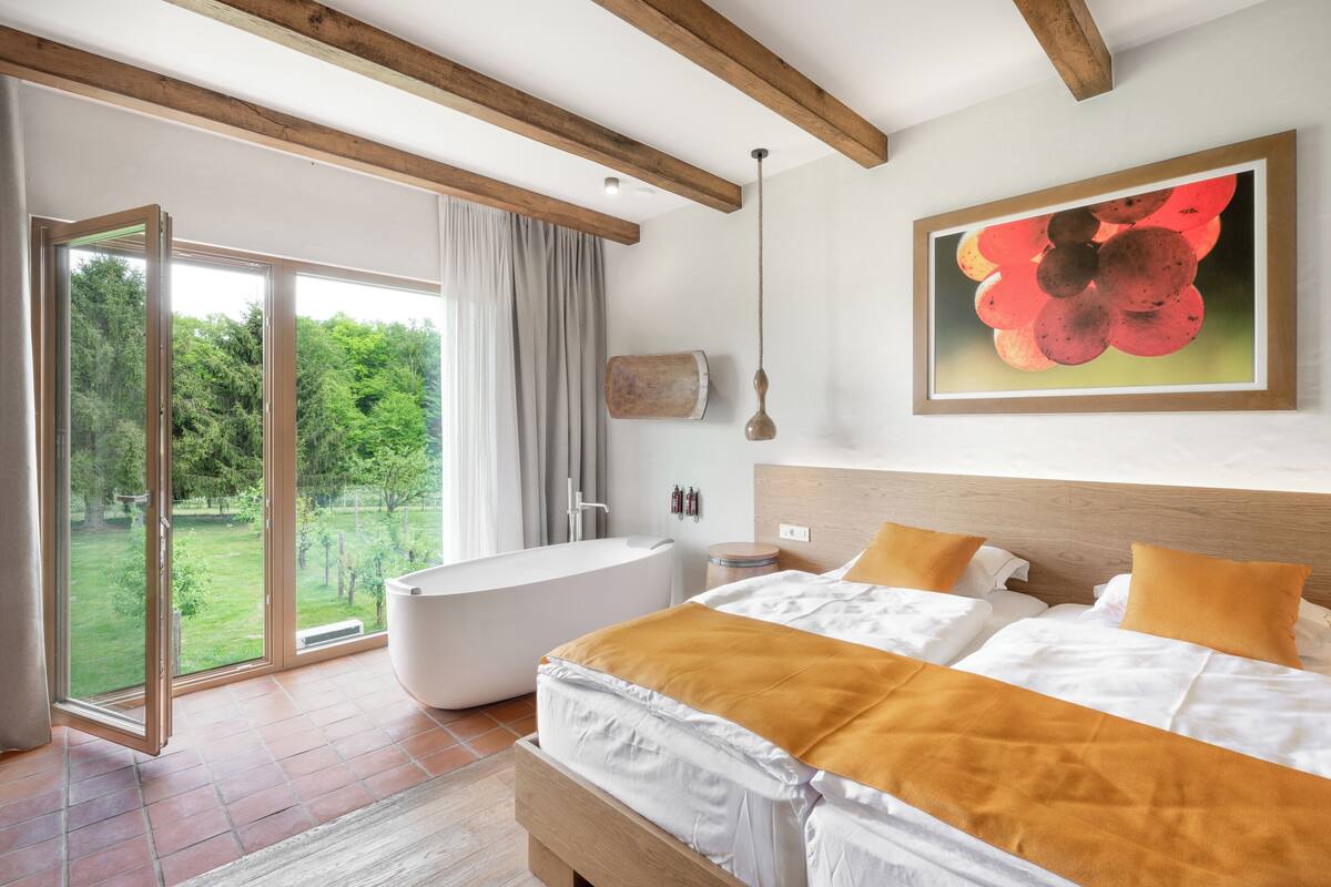 A modern bedroom with a large bed, a bathtub, and a sliding glass door leading to a garden.