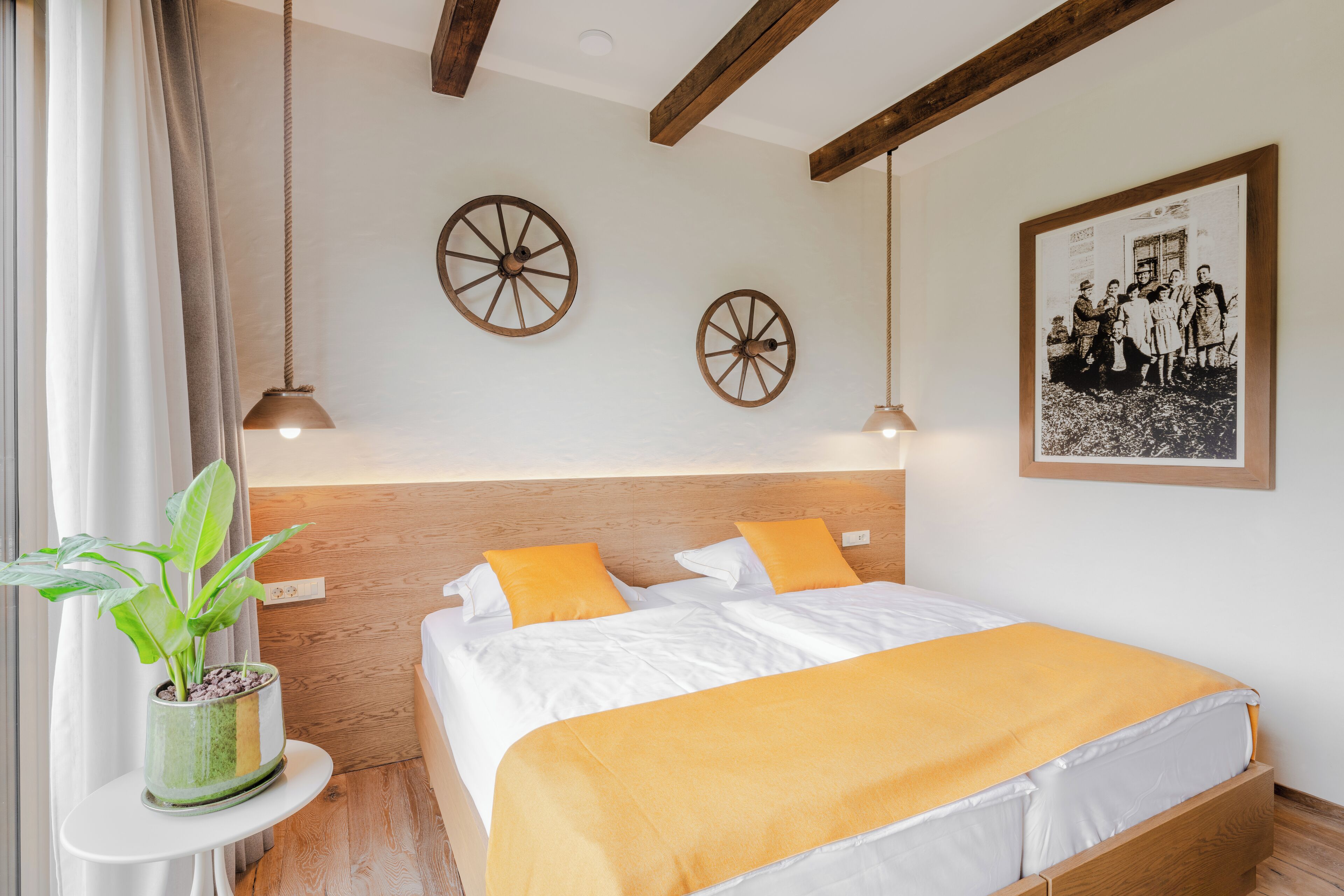 A neatly arranged bedroom with a wooden headboard, two decorative wheels on the wall, a framed family photo, and a potted plant on a side table.