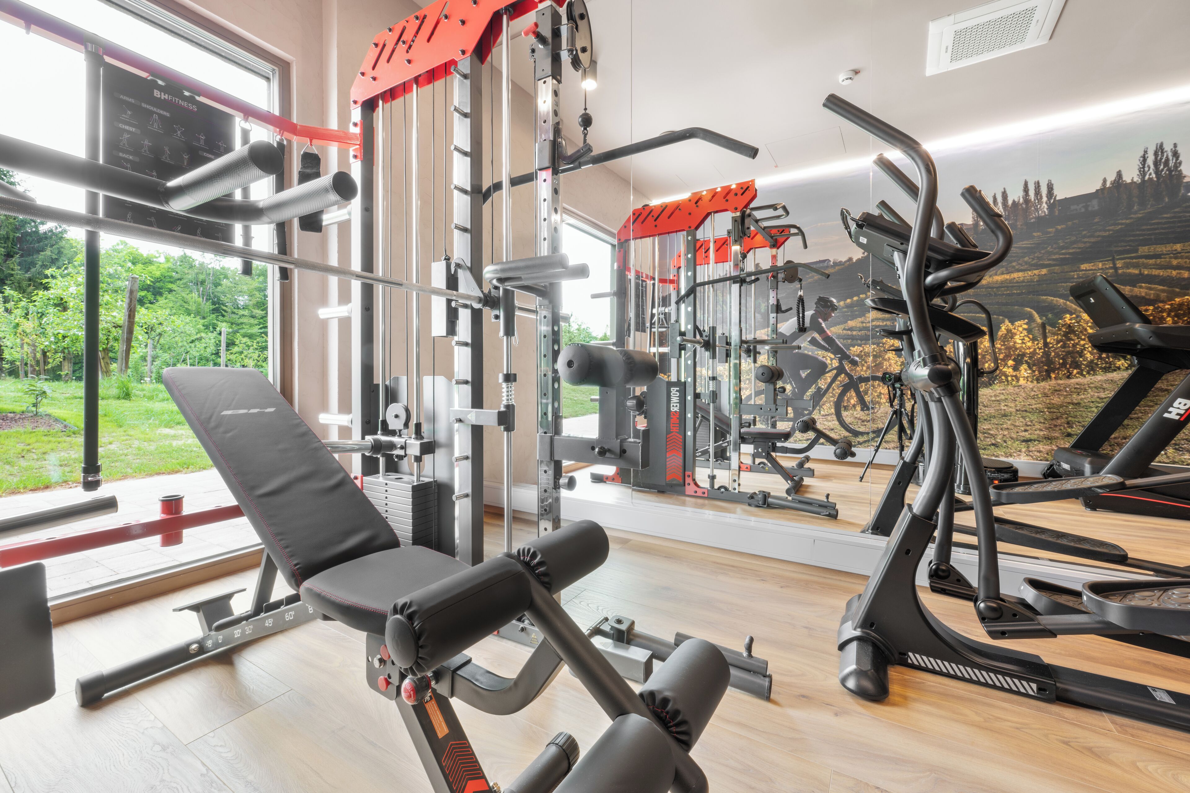A gym with exercise equipment, a bench, and a view of greenery through large windows.