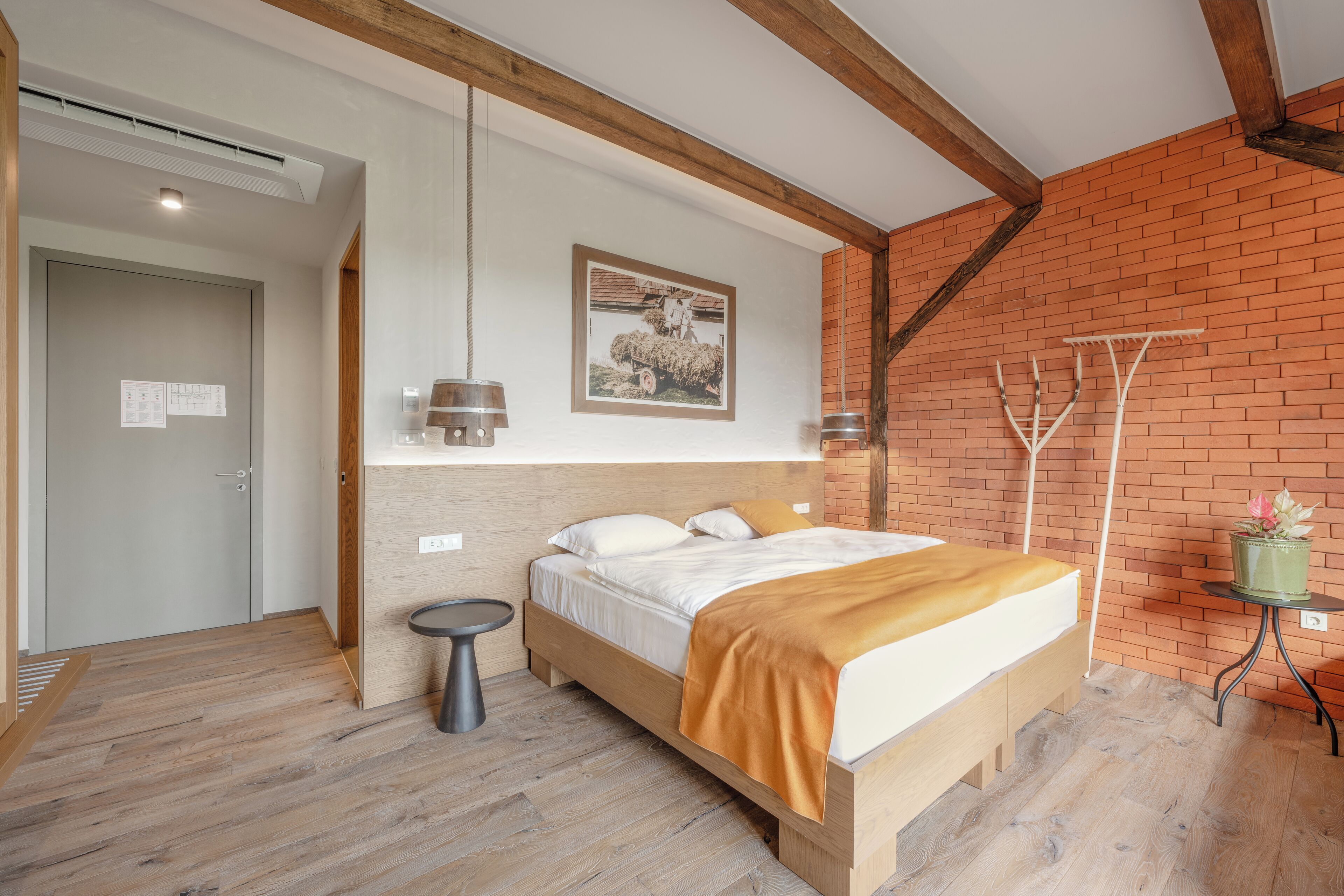 A bedroom with a wooden bed, a small round table, a wall-mounted light, and a brick wall.