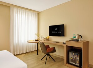 Habitación de hotel con cama, escritorio con silla, televisor, minibar y ventana con cortinas.