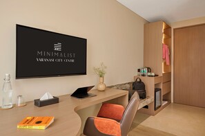 Una habitación de hotel moderna con televisión de pantalla plana, un escritorio con computadora portátil, una silla, una estantería y un armario.