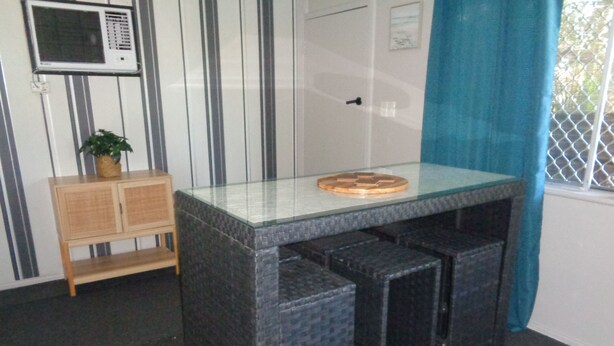 Uma pequena mesa escura com tampo de vidro e base trançada, colocada em frente a uma porta com moldura branca e um pequeno janelão com cortina azul.
