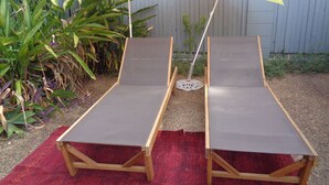Duas cadeiras de madeira com almofadas cinzas, dispostas sobre um tapete vermelho, sob dois guarda-sóis listrados.