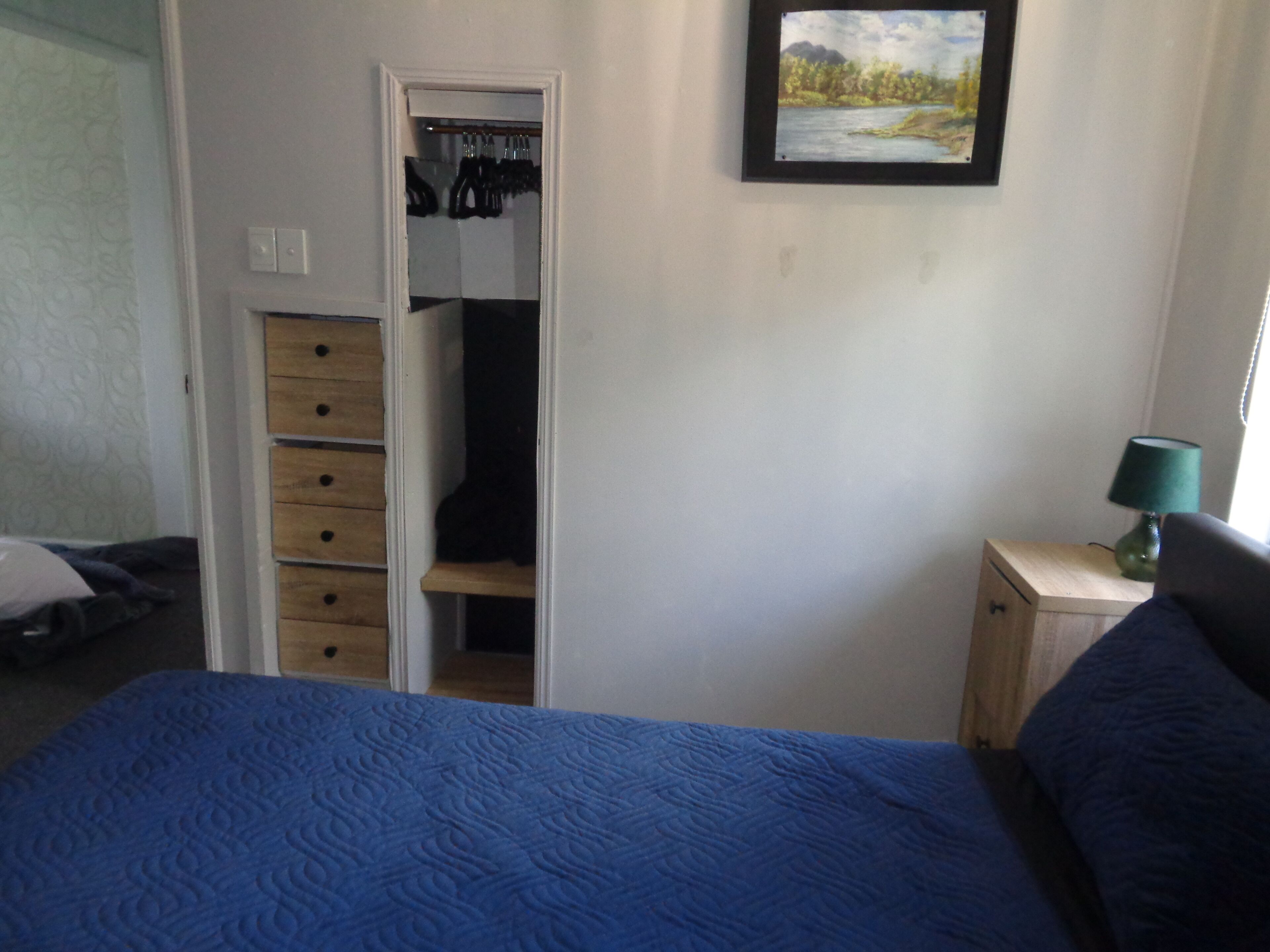 Een slaapkamer met een bed, een kledingkast, een nachtkastje met een lamp en een schilderij aan de muur.