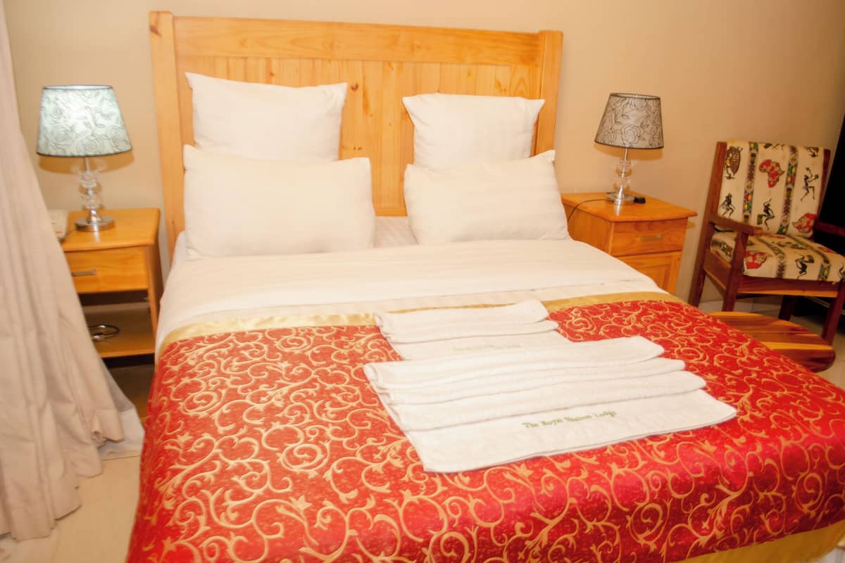 Een hotelkamer met een houten hoofdeinde, wit bedgoed, een rood beddegoed met patroon en twee nachtkastjes met lampen.