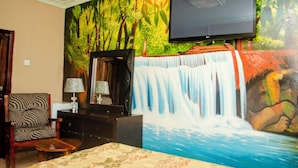 A hotel room with a large mural of a waterfall, a flat-screen TV mounted on the wall, a wooden chair, a small table, and a bed with a floral bedspread. - Royal Shalom Lodge (Lusaka)