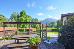 En udendørs terrasse med et træbord og bænke, omgivet af grøn vegetation og udsigt til et bjerg.