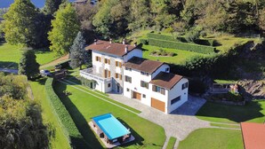 Una casa grande de varios pisos con piscina, rodeada de jardines cuidados y un paisaje ondulado.