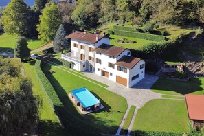 A large, multi-story house with a swimming pool, surrounded by well-maintained gardens and a hilly landscape.